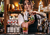 Young tattooed woman in uniform spraying finishing infusion on top of strawberry mojito cocktail during work in bar