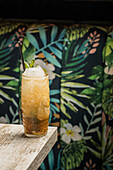 Tiki-Becher mit kaltem alkoholischem Getränk mit Strohhalm, serviert mit Eis und dekoriert mit frischen Kräutern auf unscharfem Hintergrund