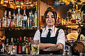 Self assured young female in uniform crossing arms and looking at camera while standing behind counter in pub