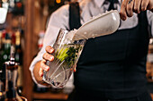 Crop female bartender in uniform pouring ice into glass while working at counter in bar