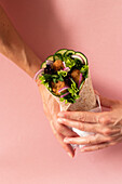 Cropped unrecognizable person hands holding vegan falafel wrap on colorful pink background
