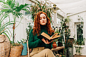 From below of concentrated red haired female sitting on chair with crossed legs and reading book among different potted plants in domestic garden