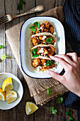 From above hand with cauliflower snack tacos in bowl and lemon pieces in plate on wooden table