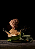 Crop anonymous person throwing tasty sweet cookies into green ceramic cup with splashing coffee on table in kitchen on black background