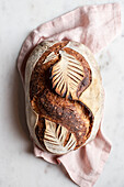 Top view of freshly baked loaf of bread with leaf ornament placed on napkin on white background in kitchen