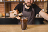 Crop bearded male barista in apron pouring syrup from metal jigger into glass of fresh iced coffee at counter