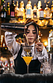 Skilled young female bartender using strainer and pouring orange alcohol drink from shaker into glass while preparing refreshing cocktail at bar counter