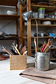 Verschiedene Töpferwerkzeuge in Metalldosen und Schreibwarenhalter auf einem Tisch in einer alten Werkstatt mit Holzregalen