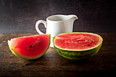 Scheiben einer reifen, süßen Wassermelone auf einem Holztisch vor dunklem Hintergrund neben einer Keramikkanne
