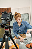 Young female blogger pouring milk from jug into cup against photo camera at table with croissants at home