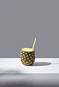 Reife Ananas mit gestreiftem Strohhalm auf einem sonnenbeschienenen Tisch, der ein frisches, gesundes Getränk auf grauem Hintergrund symbolisiert