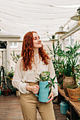 Charming female with long ginger hair standing with jug of seedling in indoor garden with various potted houseplants at home