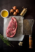 Top view of uncooked meat piece with seasonings between fresh rosemary sprigs and garlic cloves on dark background