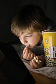 Little boy lying down comfortable on sofa with popcorn bucket while eating and watching cartoon on tablet