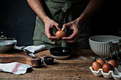 Unrecognizable guy in apron breaking raw egg over bowl while preparing pastry on lumber table near kitchenware