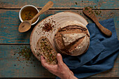 Draufsicht auf eine abgeschnittene, nicht erkennbare Hand, die eine Scheibe und einen Laib gebackenes Brot hält, das auf einem runden hölzernen Schneidebrett mit Aromastoffen in einer hellen Küche serviert wird