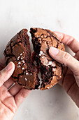 Top view of crop anonymous person breaking freshly baked chocolate brownie cookie with cracks on white background in light kitchen