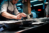 Cook holding spoon and little pan while adding sauce in plate on restaurant kitchen