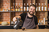 Cheerful male barista pouring hot aromatic Americano from stove top coffee maker into glass cup placed on wooden counter in cafe