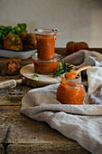 Full glass jars with homemade natural fried tomato sauce placed on cutting board board near with fresh green rosemary and basil leaves placed on rustic wooden table