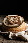 Decorative freshly baled loaf of bread with floral pattern placed on napkin on wooden table in kitchen against black background