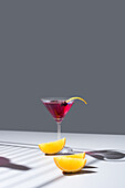 Composition of glass filled with red fruit cocktail near orange slides and zest placed on concrete table against grey background