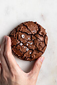 Top view of crop anonymous person breaking freshly baked chocolate brownie cookie with cracks on white background in light kitchen
