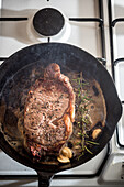 From above of pan with appetizing meat piece with condiments and rosemary sprig in melted butter on gas stove