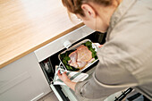 From above of crop unrecognizable female putting baking dish with uncooked poultry and spinach leaves in oven while cooking
