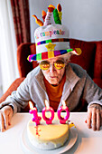 Elderly female in funny hat and sunglasses blowing burning candles with number 100 on birthday cake and celebrating anniversary at home