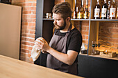 Adult bearded male barista in apron wiping moka pot with napkin while standing at counter in cafe