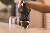 Crop anonymous male barista pouring hot aromatic Americano from stove top coffee maker into glass cup placed on wooden counter in cafe