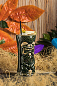 Large sculptural tiki cup filled with booze decorated with straw and fruits placed on green rug against dry grass