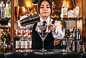 Skilled young female bartender using strainer and pouring orange alcohol drink from shaker into glass while preparing refreshing cocktail at bar counter