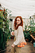 Charming young female with long wavy red hair standing with reusable string bag of tangerines in room with various houseplants