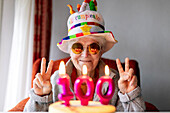 Vergnügte Seniorin mit lustigem Hut und Sonnenbrille, die mit beiden Händen das V-Zeichen zeigt und ihren Geburtstag mit einem Kuchen mit 100 Kerzen feiert