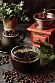 Ein Glas frisch gebrühter aromatischer Kaffee auf einer Untertasse steht auf einem Tisch mit verstreuten Bohnen vor einer manuellen Kaffeemühle