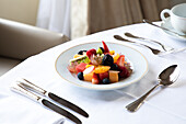 Von oben schmackhafter Obstsalat mit verschiedenen Beeren, Kiwi und Wassermelone, serviert auf einem weißen Teller auf einem Tisch mit einer Tasse Kaffee und Besteck in einem Hotelrestaurant