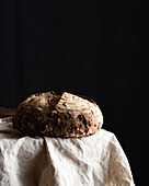 Freshly baked sourdough bread loaf with walnuts and raising placed on white linen on black background