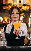 Positive young female barkeeper looking at camera and offering orange alcohol cocktail in martini glass while standing at bar counter