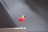 Crystal glass of pomegranate margarita cocktail served with flower blooms and leaf on concrete blocks in studio