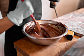 Cropped unrecognizable worker in black apron wearing gloves while melting dark chocolate in stainless steel bowl using heat gun working at counter with marble slab