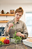 Young female with fresh green herbs in plastic box at table with peppers cooking at home