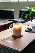 Ein Glas heißer, dampfender Tee mit Milch steht auf einem Holztisch mit einem Notizbuch in einer sonnendurchfluteten Wohnung