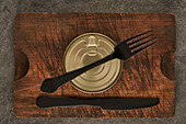 From above scratched chopping board with fork and sealed can with preserved food on rustic lumber table