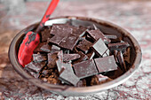 Nahaufnahme einer Edelstahlschüssel, gefüllt mit Stücken dunkler Schokolade und einem Spatel vor dem Temperieren auf einer Marmorplatte
