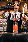 Skilled young female bartender using strainer and pouring red alcohol drink from shaker into glass with ice cubes while preparing refreshing cocktail at bar counter