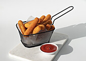Mozzarella cheese fried sticks placed in metal fryer near ketchup sauce in marble board on white background