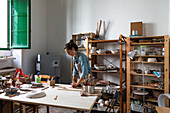 Focused adult lady in casual clothes and apron sculpting clay piece and smiling during pottery class in workshop