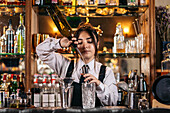 Young female barkeeper pouring alcohol drink from bottle into jigger while preparing cocktail at counter in bar